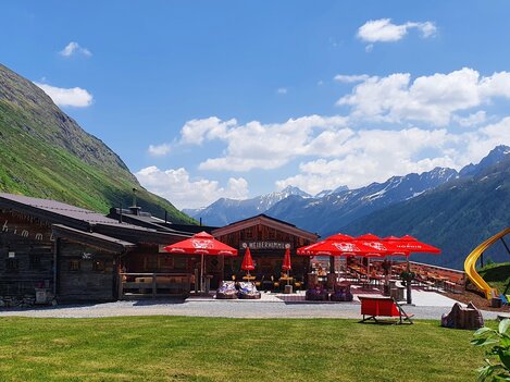 A cozy inn in the mountains with red sun umbrellas. In the background, green meadows and majestic mountains can be seen.