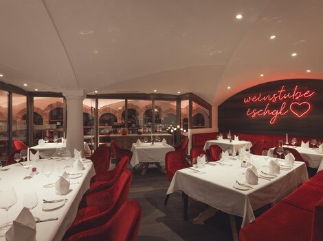 Ein stilvoller, warm beleuchteter Weinkeller mit elegant gedeckten Tischen, roten Samtsesseln und einem leuchtenden Neon-Schriftzug „Weinstube Ischgl“ schafft eine gemütlich-exklusive Atmosphäre. | © Hannes Niederkofler
