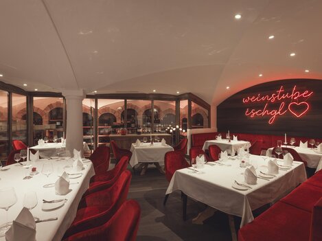 A stylish, warmly lit wine cellar with elegantly set tables, red velvet armchairs, and a glowing neon sign "Weinstube Ischgl" creates a cozy-exclusive atmosphere. | © Hannes Niederkofler