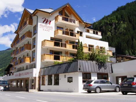 A modern hotel building with several floors and balconies. Surrounded by green mountains and clear sky.