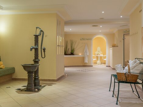 Ein eleganter Wellnessbereich mit sanften Farben und gemütlichen Möbeln. Im Mittelpunkt steht ein antiker Brunnen und eine entspannende Atmosphäre.