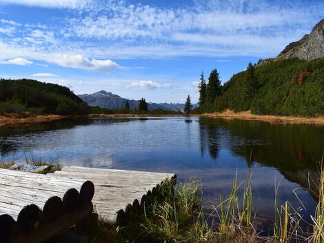 Ein ruhiger See umgeben von Bergen und Wald. Der Himmel ist klar mit einigen Wolken.