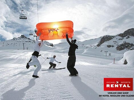 Eine Gruppe von Skifahrern steht im Schnee und hält einen orangen Schirm über sich. Im Hintergrund sind schneebedeckte Berge und ein Sessellift zu sehen.