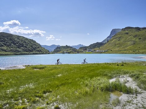 Zwei Radfahrer fahren entlang eines glitzernden Sees. Im Hintergrund sind grüne Hügel und ein blauer Himmel zu sehen.