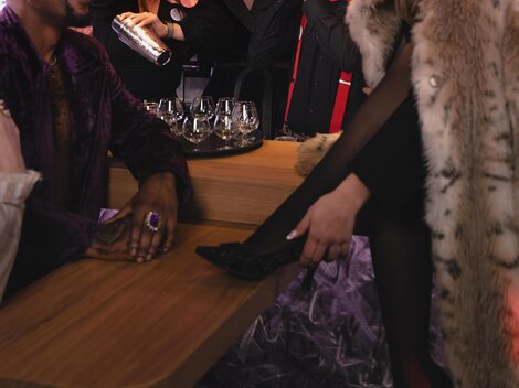 Eine lebhafte Bar-Szene mit fröhlichen Menschen, die Cocktails mixen und eine elegante Frau im Vordergrund, die entspannt sitzt. Die Atmosphäre ist glamourös und modern, beleuchtet von bunten Lichtern.