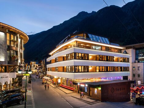 Ein modernes Gebäude in einer malerischen Alpenstadt bei Dämmerung. Im Hintergrund sind hohe Berge und eine belebte Straße mit Lichtern zu sehen.