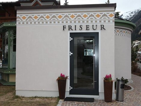 Ein Friseursalon mit einer eleganten Fassade. Die Eingangstür ist modern und es gibt Pflanzen auf beiden Seiten.