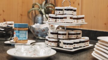 A selection of jams in small jars on a stand. Next to it is a jar of peanut butter and some plates.