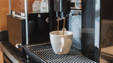 A modern coffee machine pouring fresh coffee into a white cup. In the background, some cups and the machine itself are visible.