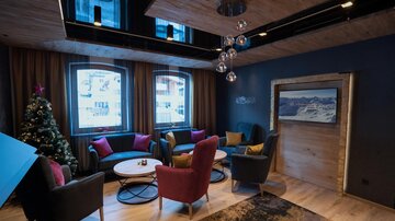 A cozy lounge with modern furniture and warm colors. In the room, there is a Christmas tree, and large windows let in plenty of daylight.