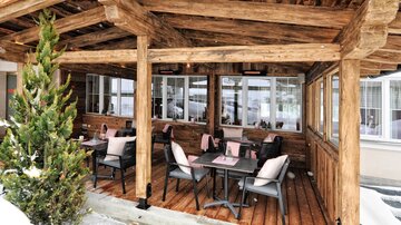 A cozy outdoor area of a restaurant with wooden construction. The tables are set, and there are seats with cushions.
