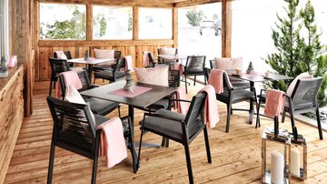 A cozy terrace with wooden paneling and modern tables. The chairs are decorated with soft cushions and there are plants in the corner.