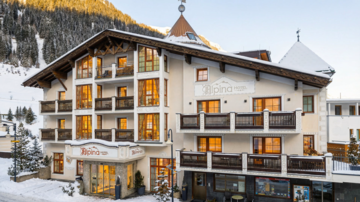 A charming hotel in Alpine style, surrounded by snow-capped mountains. Large windows allow warm light to flood the interior.