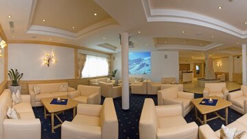 Eine elegante Lounge mit bequemen weißen Sofas und stilvollen Tischen. An der Wand ist eine Präsentation mit Berglandschaft zu sehen.