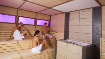 Eine moderne Sauna mit Holzbank und sanftem, farbigem Licht. Drei Menschen entspannen in handtuchähnlicher Bekleidung.