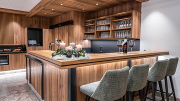 Eine moderne Küche mit einer Holzbar und eleganten Barstühlen. Auf der Theke stehen Weingläser und Dekorationen für eine einladende Atmosphäre.