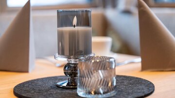 Ein eleganter Tisch mit einer Kerze in einem Glas und einem dekorativen Becher. Im Hintergrund sind graue Servietten und eine gemütliche Atmosphäre zu sehen.