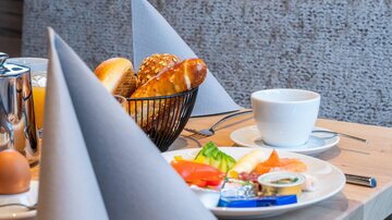 Ein gedeckter Frühstückstisch mit frischen Brötchen, einer Tasse Kaffee und frischem Obst. Im Hintergrund sind dekorierte Servietten zu sehen.