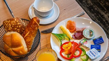 Ein Frühstückstisch mit frischen Brötchen, Aufschnitt, Käse und buntem Gemüse. Dazu ein Glas Orangensaft und eine leere Tasse.