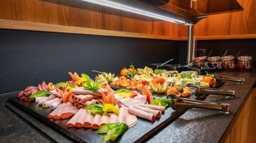 Ein reichhaltiges Buffet mit verschiedenen Wurst- und Käseplatten, garniert mit frischem Gemüse und Früchten. Die Präsentation ist ansprechend auf einem dunklen Tisch angerichtet.