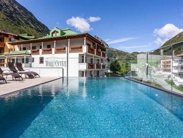 Ein modernes Hotel mit einem schönen Pool und Liegen. Im Hintergrund sind grüne Berge und ein klarer Himmel zu sehen.