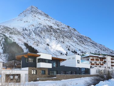 Ein modernes Gebäude im Vordergrund, umgeben von schneebedeckten Bergen. Der klare, blaue Himmel vervollständigt die winterliche Landschaft.