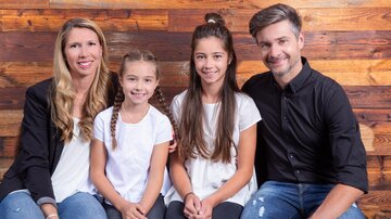 A family poses in front of a wood paneling. Two girls sit between their parents, all smiling kindly.
