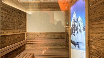 A modern room with wooden elements and a sauna bench. On the wall, there is a large screen image of snow-covered mountains.