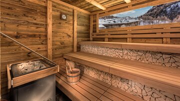 A cozy sauna with wooden benches and a stone oven. A wintry mountain landscape is visible through the window.