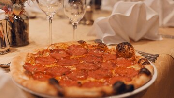 Eine leckere Pizza mit Peperoni auf einem Tisch. Im Hintergrund sind Gläser und dekorierte Servietten zu sehen.