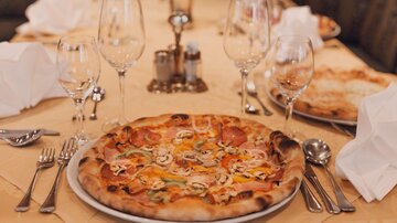 Ein gedeckter Tisch mit mehreren Gläsern und Besteck. In der Mitte liegt eine große pizza mit verschiedenen Belägen.