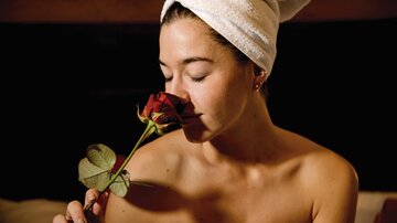 Eine Frau mit einem Handtuch auf dem Kopf riecht an einer roten Rose. Sie wirkt entspannt und zufrieden.