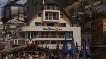 Ein modernes Hotelgebäude in den Bergen mit Terrassen und einem Seilbahnanschluss. Im Vordergrund sind Restaurant-Tische und Sonnenschirme zu sehen.
