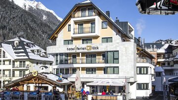 Ein modernes Hotel in den Alpen, umgeben von schneebedeckten Bergen. Vor dem Hotel gibt es eine Terrasse mit Gästen, die die Winterlandschaft genießen.
