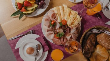 Ein ansprechend angerichteter Frühstückstisch mit Wurst, Käse und frischem Obst. Dazu gibt es verschiedene Brote und Getränke.