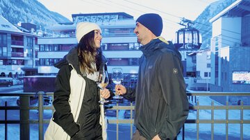 Ein Paar steht draußen und hält Gläser mit Getränken in der Hand. Im Hintergrund sind schneebedeckte Berge und eine Seilbahn zu sehen.