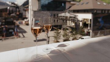 Zwei Gläser mit Rosé-Sekt stehen auf einem Tisch, mit einem Blick auf die Gebirgslandschaft und Gebäude im Hintergrund. Die Szene vermittelt ein Gefühl von Entspannung und Genuss.