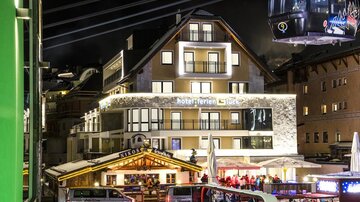 Ein modernes Hotel mit beleuchtetem Balkon in einer Stadt bei Nacht. Im Vordergrund sind Stände und Fahrzeuge zu sehen.