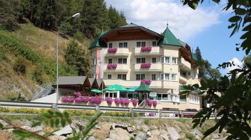 Ein charmantes Hotel mit bunten Blumenbalkonen, umgeben von Bäumen. Im Hintergrund ist ein strahlend blauer Himmel zu sehen.