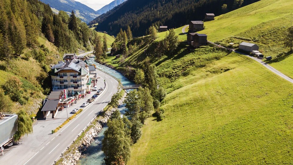 Eine malerische Landschaft mit grünen Wiesen und einem kleinen Fluss. Im Hintergrund sind Berge und einige Holzgebäude zu sehen.