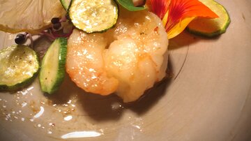 Ein elegantes Gericht mit Garnelen, Zucchini und Limettenscheiben. Mit essbaren Blüten dekoriert, auf einem grauen Teller serviert.