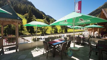 Eine gemütliche Terrasse mit Tischen und Sonnenschirmen. Im Hintergrund erstreckt sich eine malerische Berglandschaft.