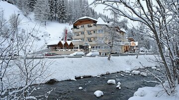 Ein malerisches winterliches Szenario mit schneebedeckten Bäumen und einem klaren Fluss. Im Hintergrund ist ein gemütliches Hotel zu sehen, umgeben von der verschneiten Landschaft.