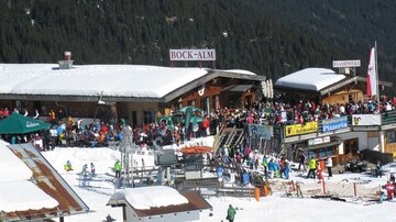 Ein belebter Skihang mit vielen Menschen. Im Vordergrund sind Skihütten und Stände in einer schneebedeckten Landschaft zu sehen.