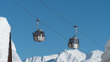 Zwei Gondelbahnen schweben über schneebedeckte Berge unter einem strahlend blauen Himmel. Die Szene vermittelt ein Gefühl von Freiheit und Abenteuer in den Bergen.