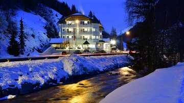 Ein malerisches Hotel in den Bergen, umgeben von Schnee und einem Fluss. Die Nachtbeleuchtung schafft eine gemütliche Atmosphäre.