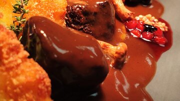 Ein elegantes Gericht mit saftigem Fleisch, dunkler Sauce und Beilagen. Dazu sind frische Kräuter und eine knusprige Komponente zu sehen.