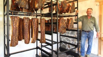 Ein Mann steht neben einem Gestell mit getrocknetem Fleisch. Im Hintergrund hängen mehrere Stücke Trockenfleisch in einer ländlichen Umgebung.
