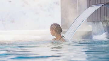 Eine Person entspannt im Wasser eines Außenpools. Im Hintergrund sind schneebedeckte Landschaften und Gebäude zu sehen.