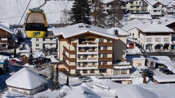 Eine Bergstation in einer schneebedeckten Landschaft mit einem Skilift. Im Vordergrund sind mehrere Gebäude und parkende Autos sichtbar.